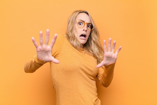 Joven mujer bonita rubia sintiéndose aterrorizada, retrocediendo y gritando de horror y pánico, reaccionando a una pesadilla contra la pared de color plano