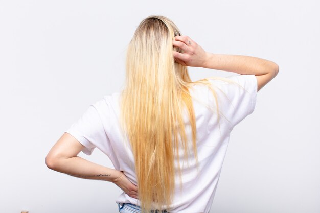 Joven mujer bonita rubia sintiéndose desorientada y confundida, pensando en una solución, con la mano en la cadera y otra en la cabeza, vista posterior sobre la pared blanca