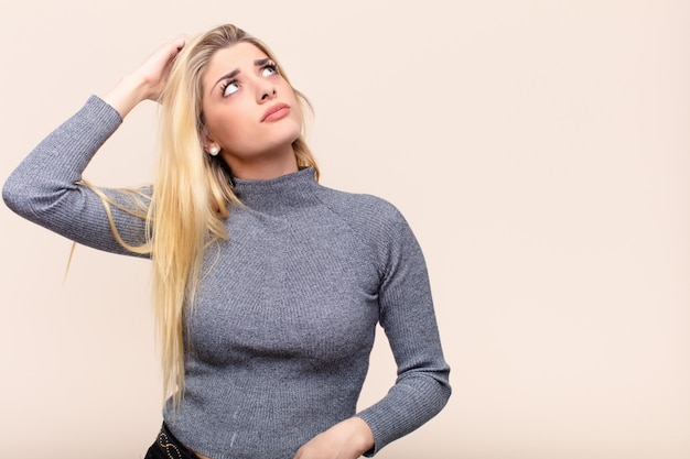 Joven mujer bonita rubia sintiéndose perpleja y confundida, rascándose la cabeza y mirando hacia un lado sobre la pared