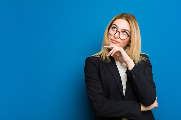 Joven mujer bonita rubia sintiéndose reflexiva, preguntándose o imaginando ideas, soñando despierta y mirando hacia arriba para copiar el espacio contra la pared plana