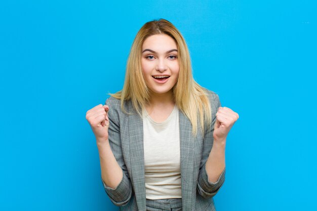 Joven mujer bonita rubia sintiéndose sorprendida, emocionada y feliz, riendo y celebrando el éxito, diciendo wow! contra pared plana