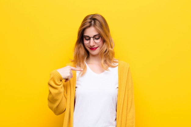 Joven mujer bonita rubia sonriendo alegremente y casualmente, mirando hacia abajo y apuntando al pecho contra la pared amarilla