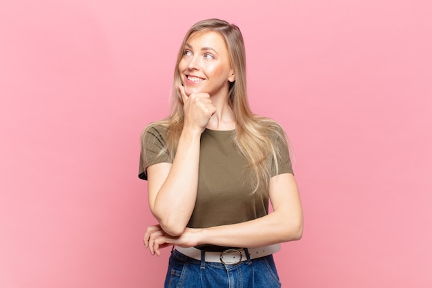 Joven mujer bonita rubia sonriendo con una expresión feliz y segura con la mano en la barbilla, preguntándose y mirando hacia un lado