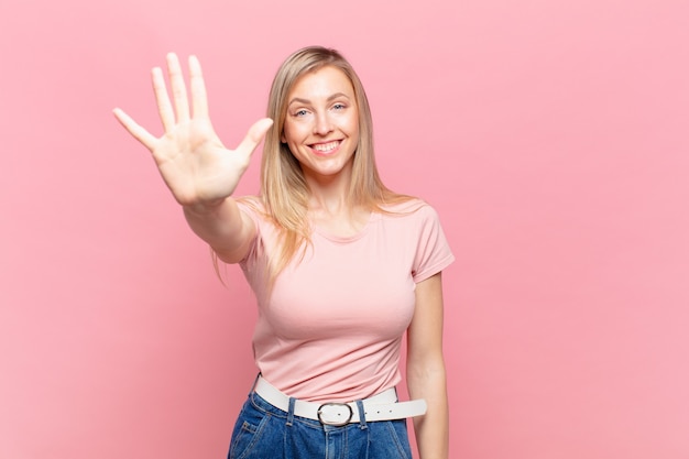 Joven mujer bonita rubia sonriendo y mirando amistosamente, mostrando el número cinco o quinto con la mano hacia adelante, contando hacia atrás