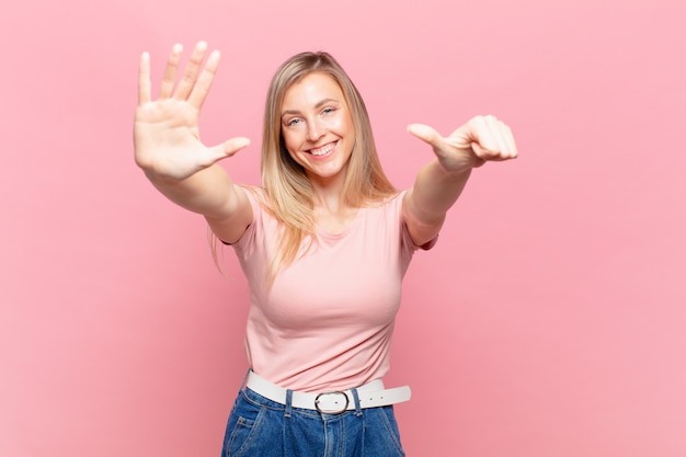 Joven mujer bonita rubia sonriendo y mirando amistosamente, mostrando el número seis o sexto con la mano hacia adelante, contando hacia atrás