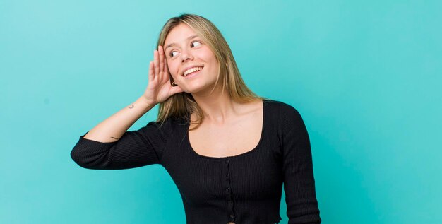 Joven mujer bonita rubia sonriendo mirando curiosamente a un lado tratando de escuchar chismes o escuchar un secreto