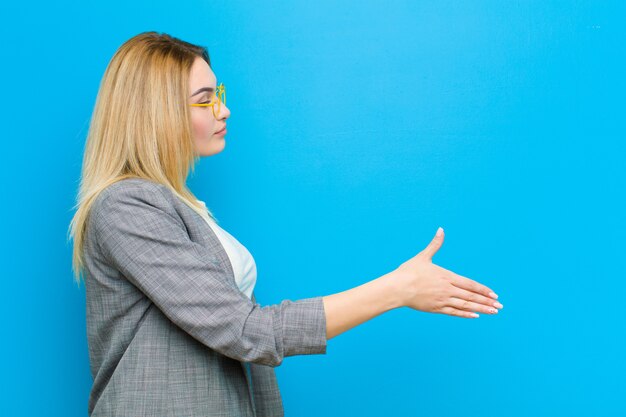 Joven mujer bonita rubia sonriendo, saludando y ofreciendo un apretón de manos para cerrar un acuerdo exitoso, concepto de cooperación contra la pared plana