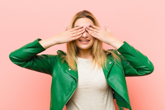 Foto joven mujer bonita rubia sonriendo y sintiéndose feliz, cubriendo los ojos con ambas manos y esperando una sorpresa increíble sobre una pared plana rosa