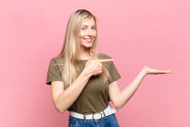 Joven mujer bonita rubia sonriendo, sintiéndose feliz, despreocupada y satisfecha, apuntando al concepto o idea en el espacio de la copia en el lateral