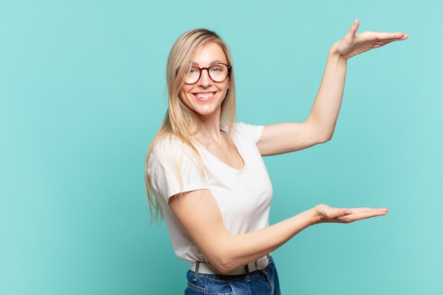 Joven mujer bonita rubia sonriendo, sintiéndose feliz, positiva y satisfecha, sosteniendo o mostrando un objeto o concepto en el espacio de la copia