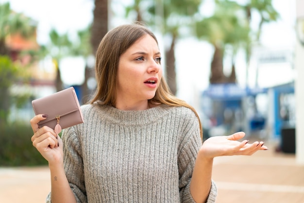 Joven mujer bonita rubia sosteniendo una billetera al aire libre con expresión facial sorpresa