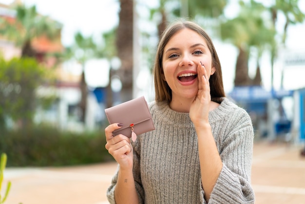 Joven mujer bonita rubia sosteniendo una billetera al aire libre gritando con la boca abierta