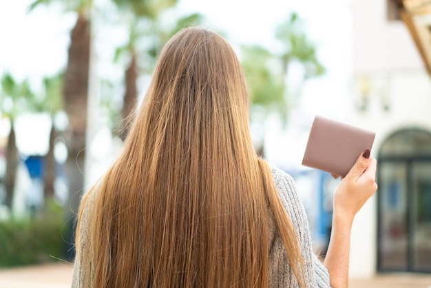 Joven mujer bonita rubia sosteniendo una billetera al aire libre en posición trasera