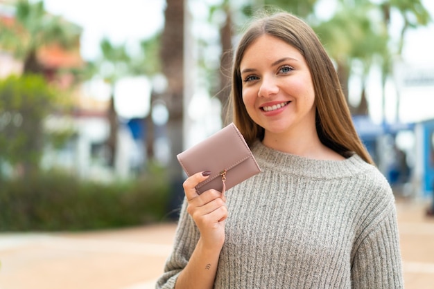 Joven mujer bonita rubia sosteniendo una billetera al aire libre sonriendo mucho