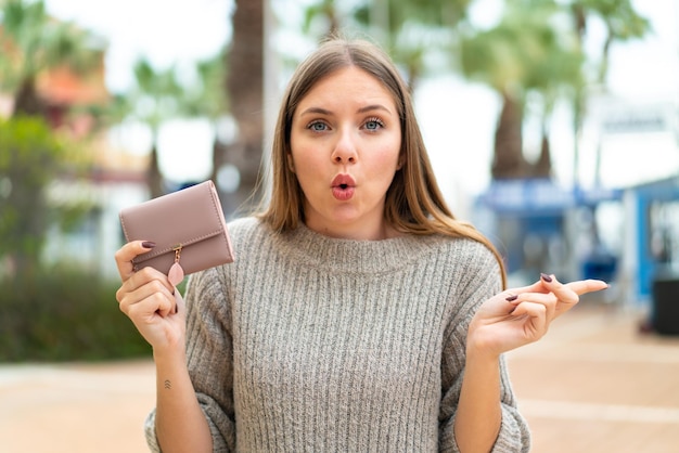 Joven mujer bonita rubia sosteniendo una billetera al aire libre sorprendida y apuntando hacia el lado