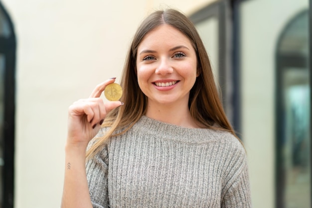 Joven mujer bonita rubia sosteniendo un Bitcoin al aire libre sonriendo mucho