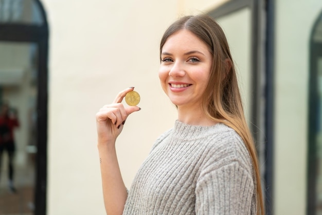 Joven mujer bonita rubia sosteniendo un Bitcoin al aire libre sonriendo mucho