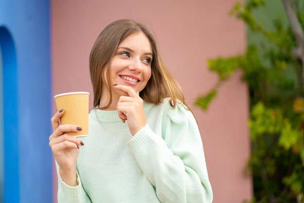 Joven mujer bonita rubia sosteniendo un café para llevar al aire libre pensando en una idea y mirando de lado