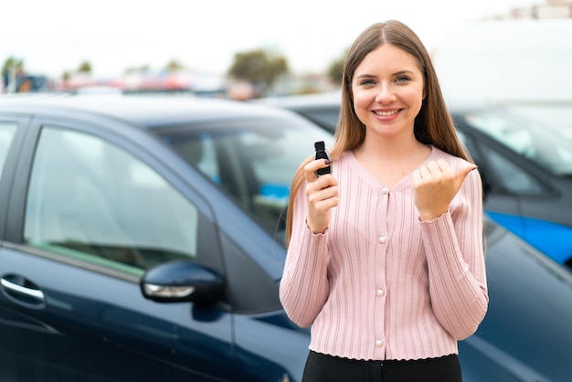 Joven mujer bonita rubia sosteniendo las llaves del auto al aire libre apuntando hacia un lado para presentar un producto