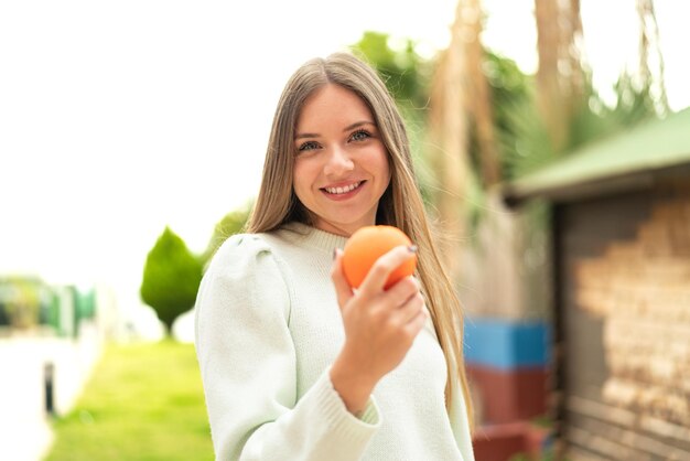 Joven mujer bonita rubia sosteniendo una naranja al aire libre con expresión feliz