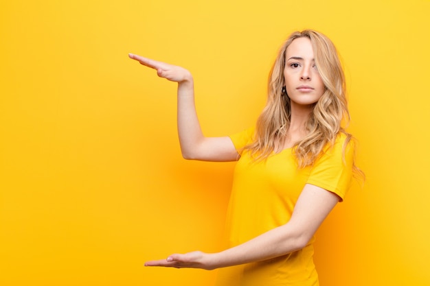 Joven mujer bonita rubia sosteniendo un objeto con ambas manos en el espacio de copia lateral, mostrando, ofreciendo o publicitando un objeto contra la pared de color plano