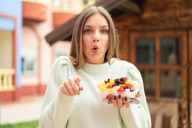 Foto joven mujer bonita rubia sosteniendo un plato de fruta al aire libre sorprendida y apuntando al frente