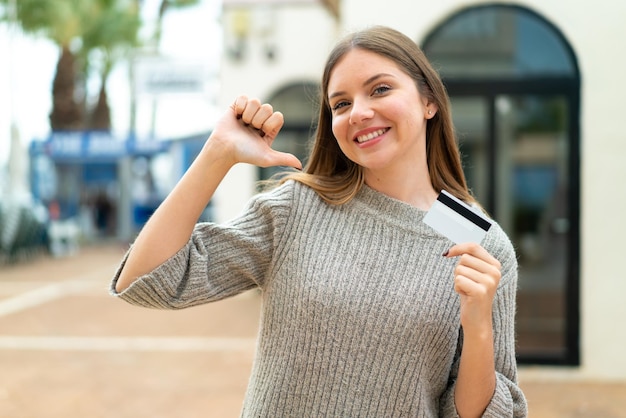 Joven mujer bonita rubia sosteniendo una tarjeta de crédito al aire libre orgullosa y satisfecha