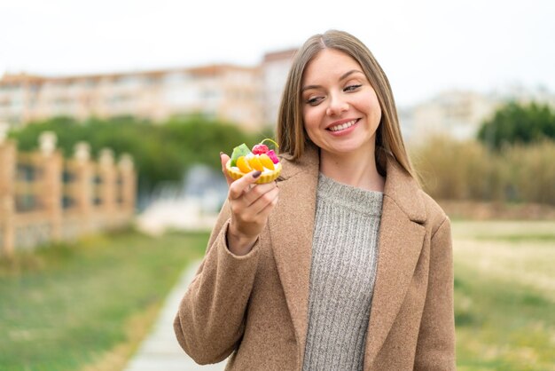 Joven mujer bonita rubia sosteniendo una tartaleta al aire libre con expresión feliz