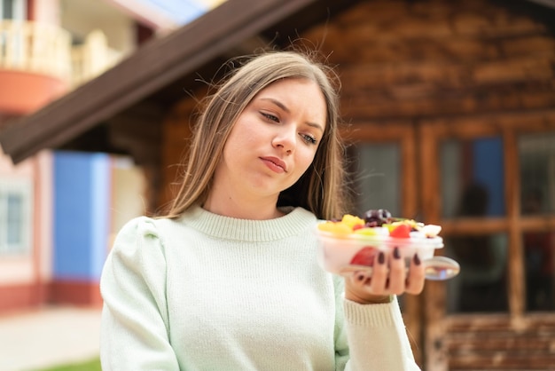 Joven mujer bonita rubia sosteniendo un tazón de fruta al aire libre con expresión triste