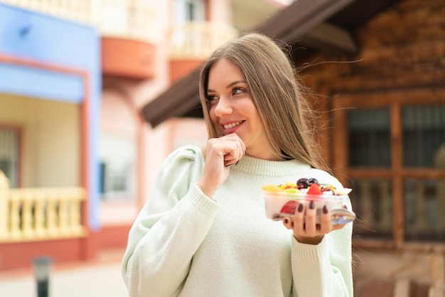 Joven mujer bonita rubia sosteniendo un tazón de fruta al aire libre pensando en una idea y mirando de lado