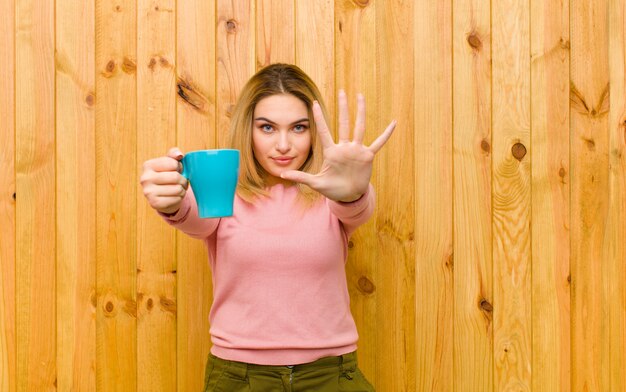 Joven mujer bonita rubia con una taza de café en la pared de madera