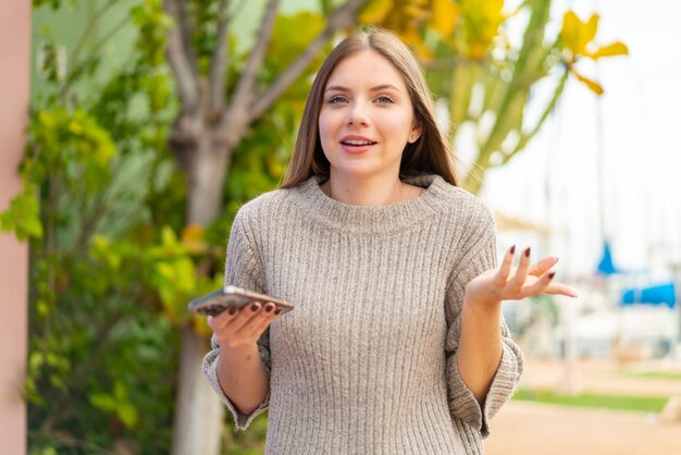 Joven mujer bonita rubia usando teléfono móvil al aire libre con expresión facial sorprendida