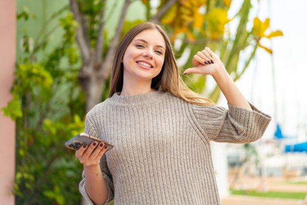 Joven mujer bonita rubia usando teléfono móvil al aire libre orgullosa y satisfecha