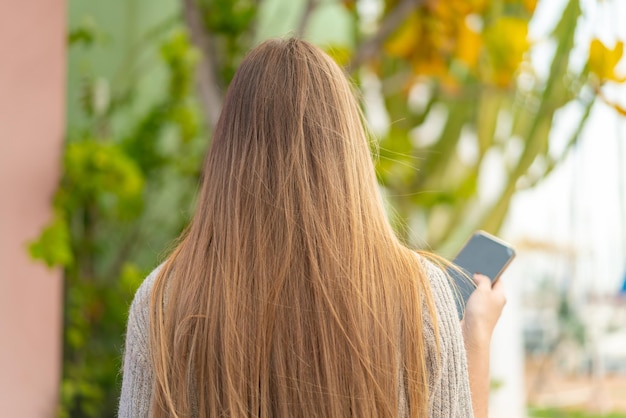 Joven mujer bonita rubia usando teléfono móvil al aire libre en posición trasera