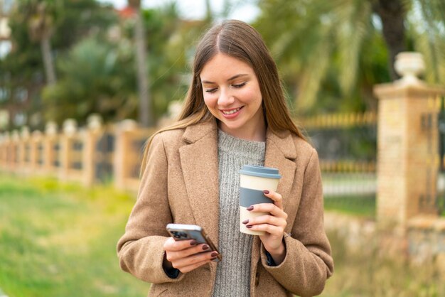 Joven mujer bonita rubia usando teléfono móvil y sosteniendo un café con expresión feliz