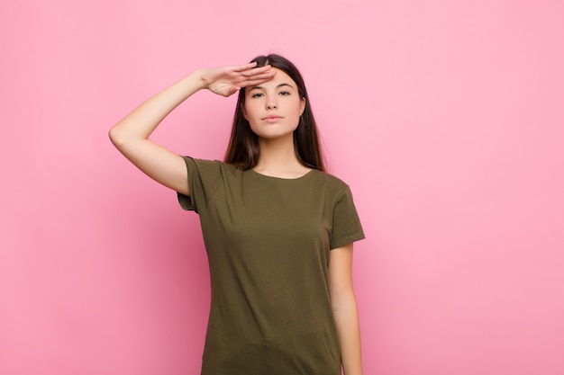 Joven mujer bonita saludando a la cámara con un saludo militar en un acto de honor y patriotismo, mostrando respeto por la pared rosa