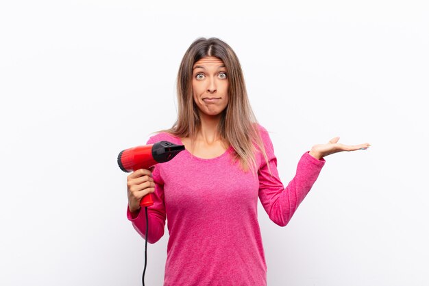 Joven mujer bonita se siente perpleja y confundida, dudando, sopesando o eligiendo diferentes opciones con expresión divertida con un secador de pelo.