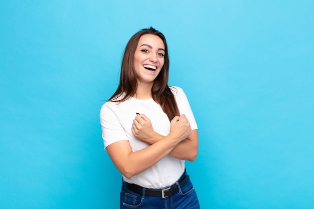Foto joven mujer bonita sonriendo alegremente y celebrando, con los puños cerrados y los brazos cruzados, sintiéndose feliz y positiva contra la pared azul