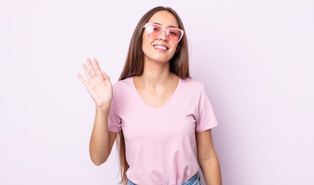 Joven mujer bonita sonriendo alegremente, saludando con la mano, dándote la bienvenida y saludándote. concepto de gafas de sol rosas