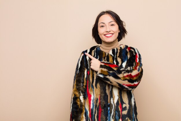 Joven mujer bonita sonriendo alegremente, sintiéndose feliz y apuntando hacia un lado y hacia arriba, mostrando el objeto en el espacio de la copia contra la pared de color beige