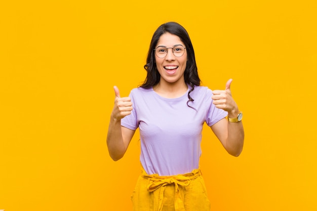 Joven mujer bonita sonriendo ampliamente mirando feliz, positiva, segura y exitosa, con ambos pulgares contra la pared naranja