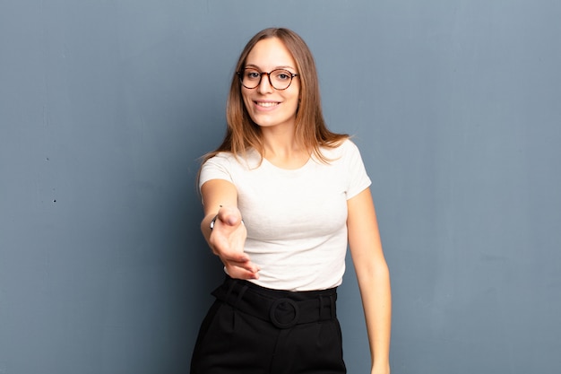 Joven mujer bonita sonriendo, luciendo feliz, confiada y amigable, ofreciendo un apretón de manos para cerrar un trato, cooperando sobre una pared gris