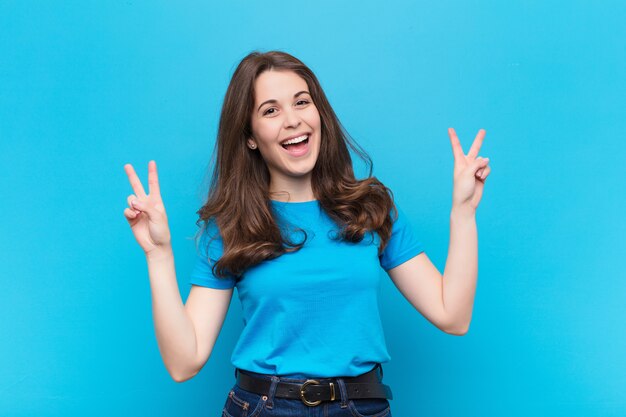 Joven mujer bonita sonriendo y mirando feliz, amigable y satisfecha, gesticulando victoria o paz con ambas manos sobre la pared azul