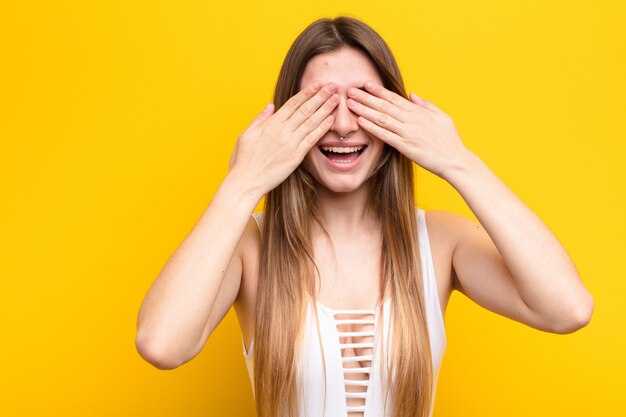 Joven mujer bonita sonriendo y sintiéndose feliz, cubriendo los ojos con ambas manos y esperando una sorpresa increíble