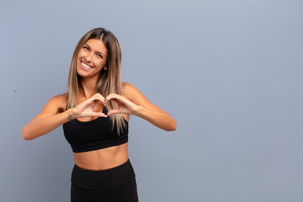 Joven mujer bonita sonriendo y sintiéndose feliz, linda, romántica y enamorada, haciendo forma de corazón con ambas manos