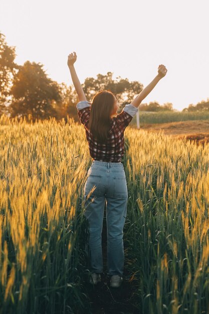 Joven mujer bonita en vestido rojo de verano y sombrero de paja caminando por el campo amarillo de la granja con trigo dorado maduro disfrutando de la cálida noche