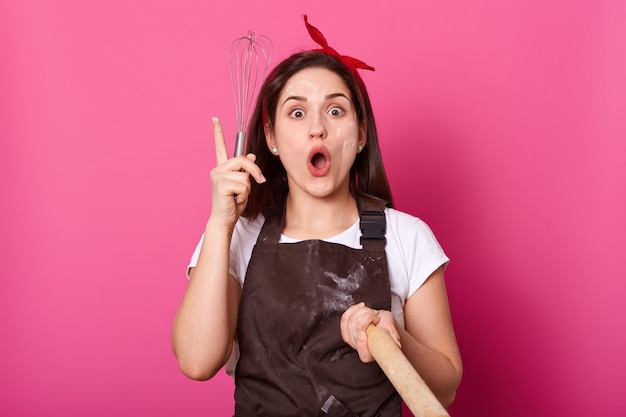 Joven mujer de cabello oscuro con delantal y cinta para el pelo roja, se para con la boca abierta, sostiene un rodillo y bate en las manos, tiene una nueva idea para hacer pastel, señala con el dedo hacia arriba en rosa. Concepto culinario