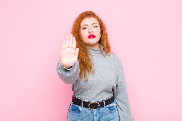 Joven mujer de cabeza roja que parece seria, severa, disgustada y enojada mostrando la palma abierta haciendo un gesto de parada contra la pared rosa