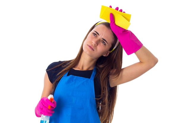 Joven mujer cansada con camiseta azul y delantal con guantes rosas sosteniendo plumero y detergente en el estudio