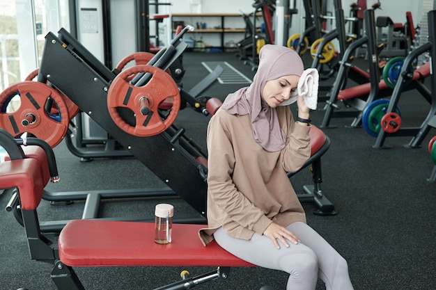 Joven mujer cansada en hijab y ropa deportiva que descansan después de un duro entrenamiento deportivo en el gimnasio